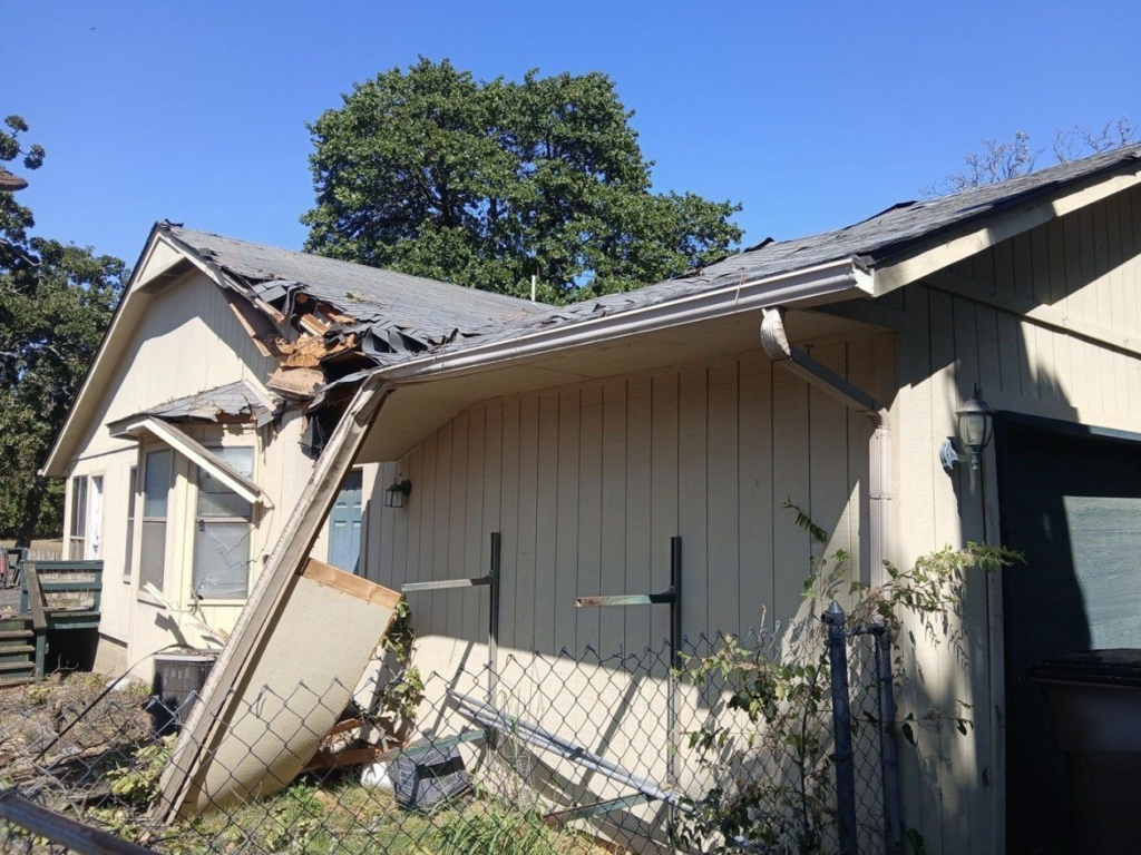 Storm-damaged roof torn open above a home's side porch in Sperry, with the porch overhang collapsed and the gutter pulled loose