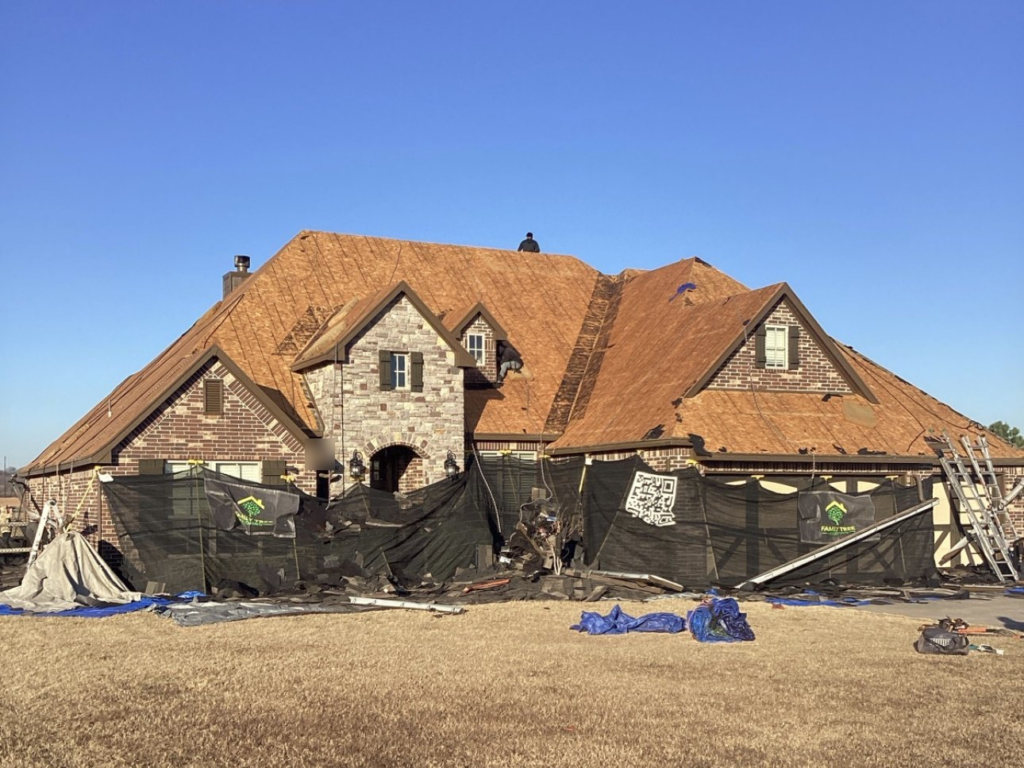 Roof torn off to the bare wood decking on a brick and stone home in Glenpool, with Catch-All netting and tarps protecting the house and yard