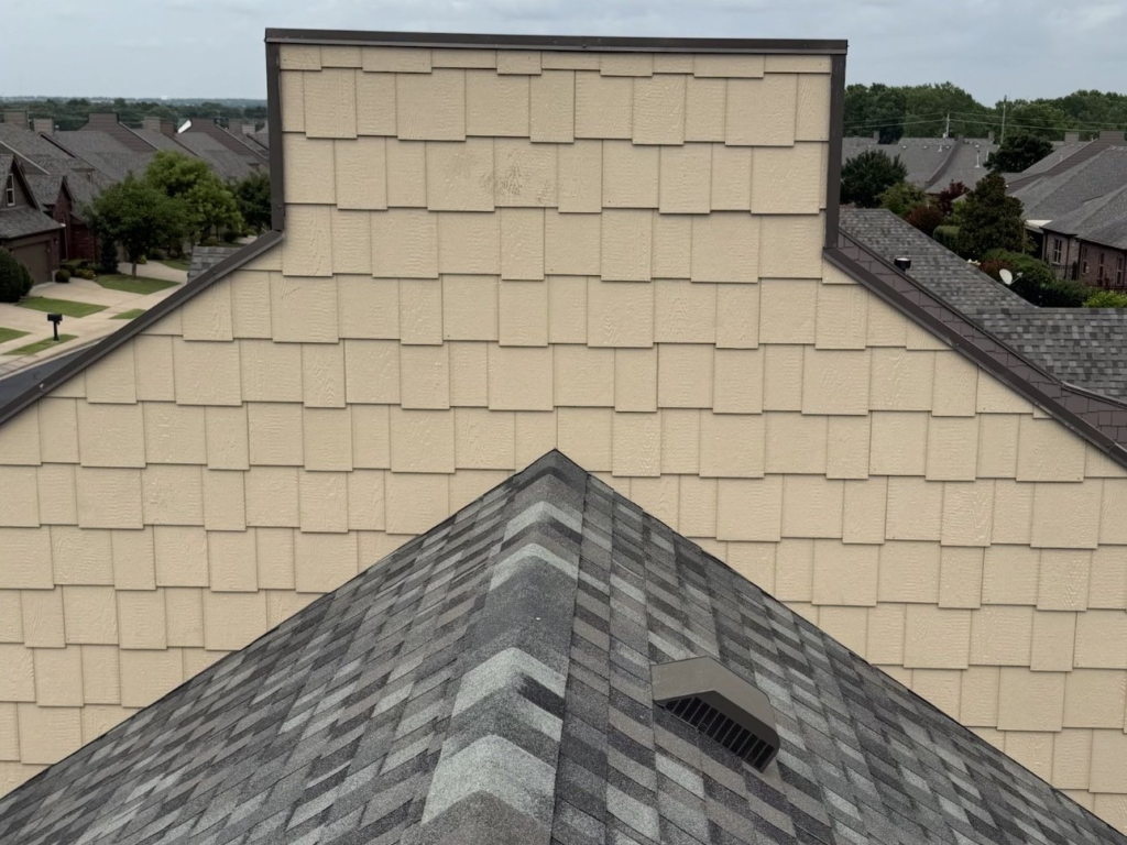 Tan shake-style siding on a gable wall above a gray shingle roof with box vents